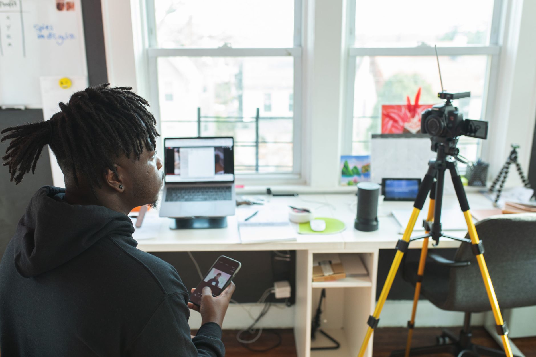 Young man using a smartphone and camera setup in a bright home office. Productive and tech-savvy environment.