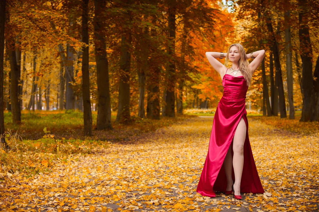 a woman in a red dress standing on a path covered with autumn leaves