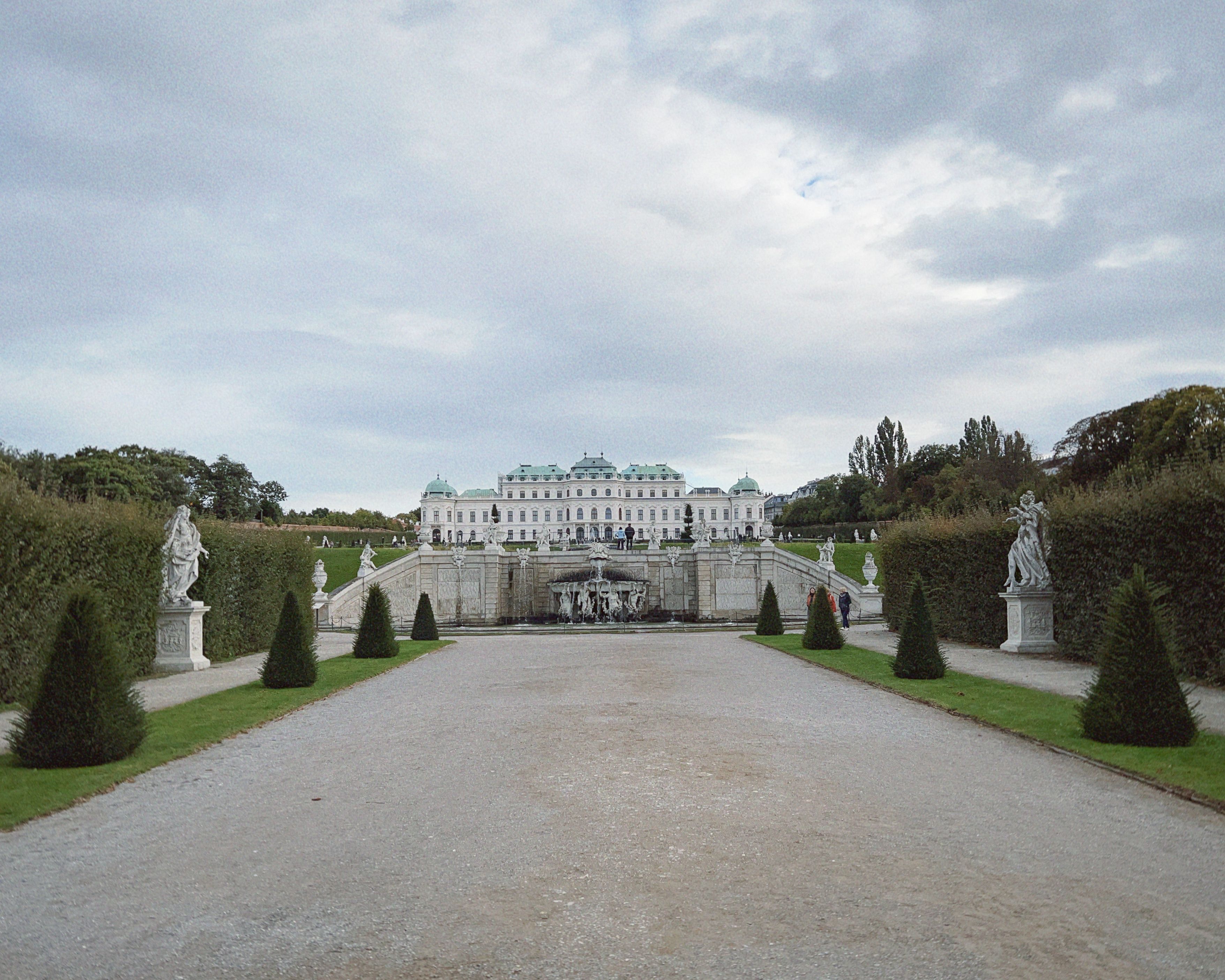 View of a grand baroque palace with symmetric formal gardens under a cloudy sky.