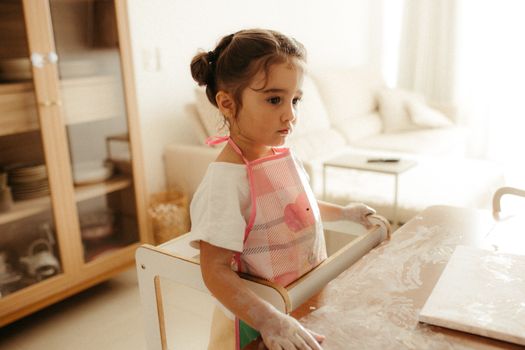 A young girl in an apron explores creativity in the kitchen by playing with flour on a table.
