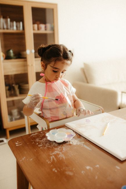 A young child painting indoors with watercolors, wearing a protective apron at a wooden table.