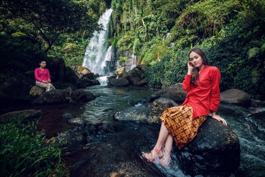 Two women in traditional attire enjoy a peaceful moment by a waterfall in Jawa Barat, Indonesia.