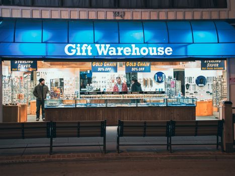 Night view of Gift Warehouse store with vibrant blue signage and two people inside.