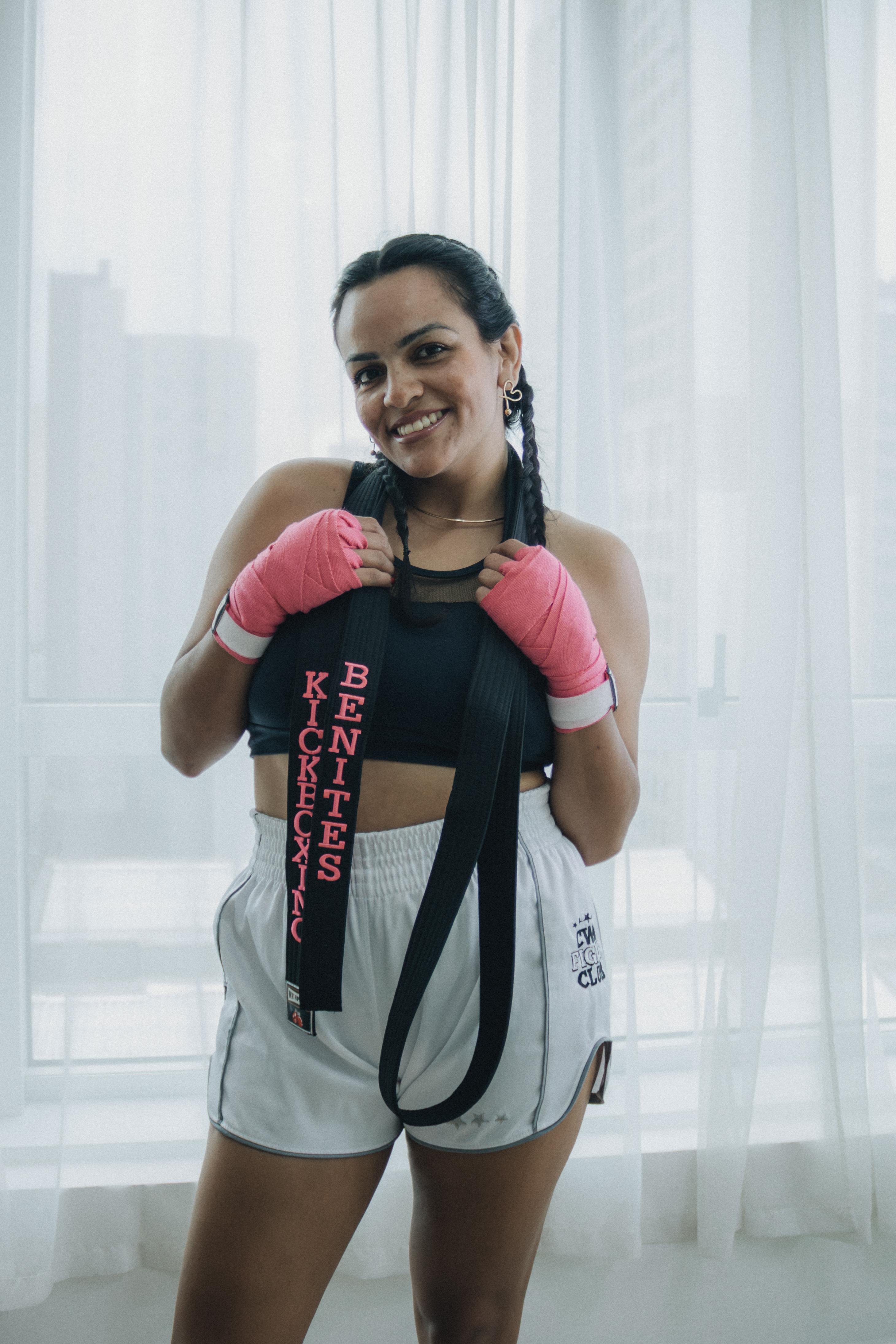 Smiling female kickboxer in pink gloves holding a championship belt indoors.