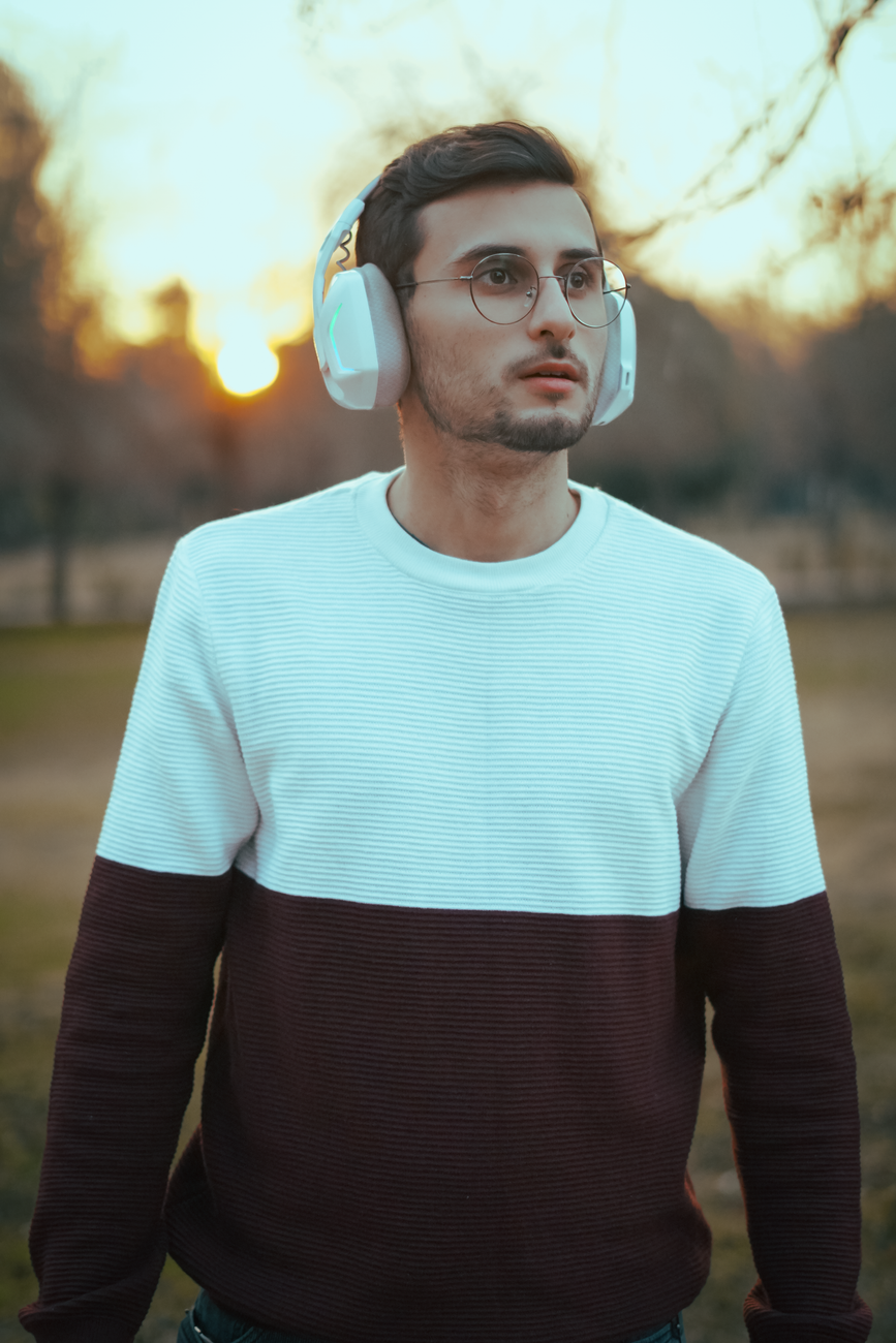 Stylish young man at sunset in park wearing glasses and headphones, enjoying music outdoors.