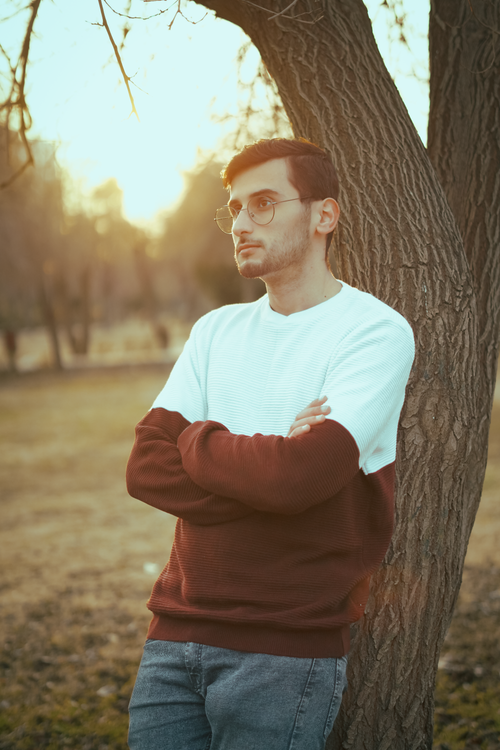Free Casually dressed young man with glasses leaning on a tree in a park during sunset. Stock Photo