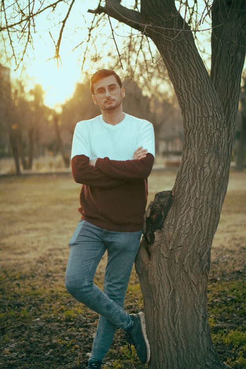 Free Casual young man in stylish outfit leaning against a tree at sunset in a park. Stock Photo