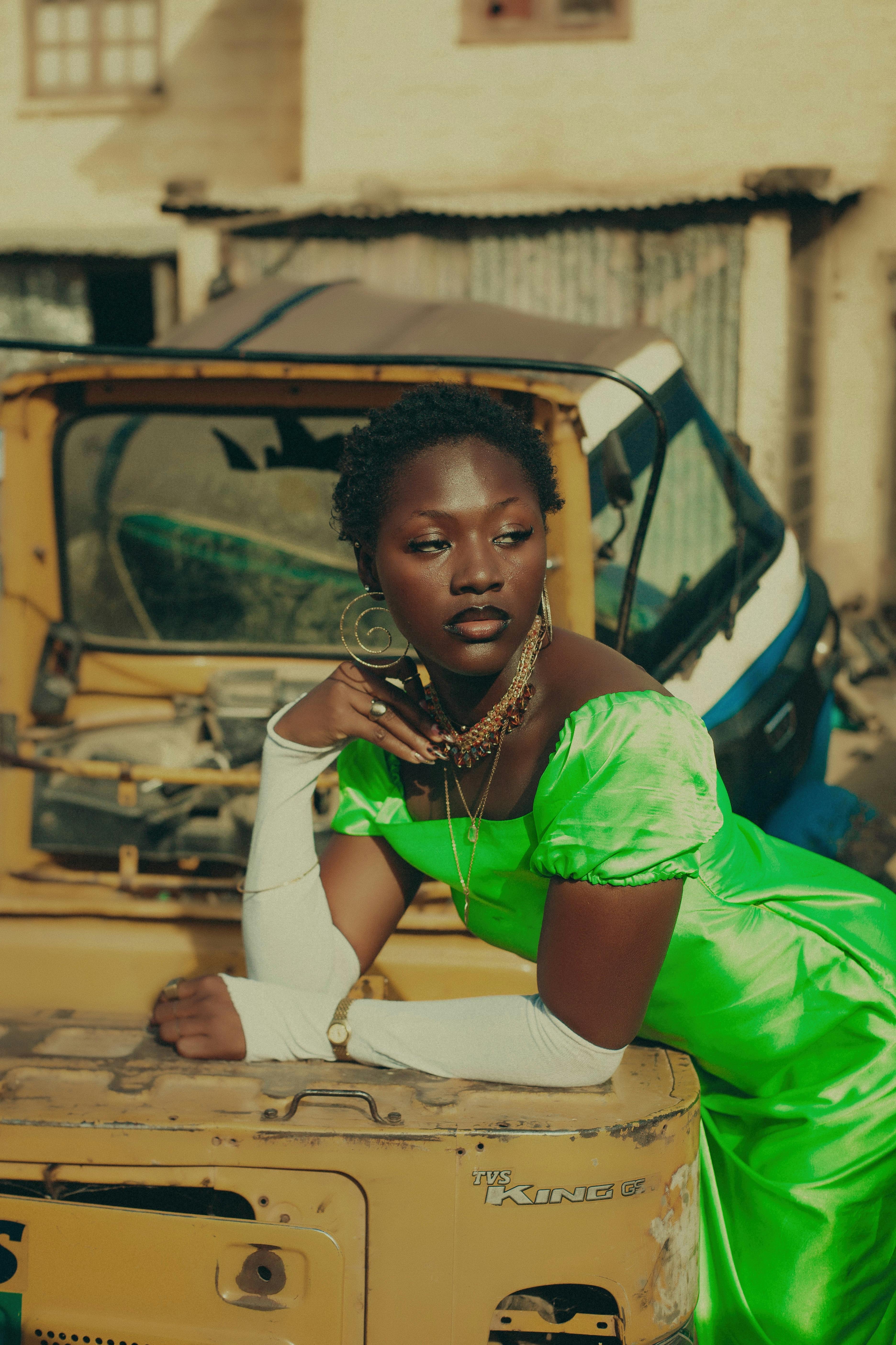 Stylish woman in vibrant green dress leans on a vintage vehicle outdoors.
