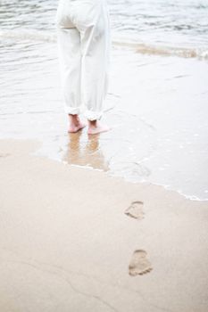 Close-up of a woman’s legs standing by the ocean, footprints leading to the water.