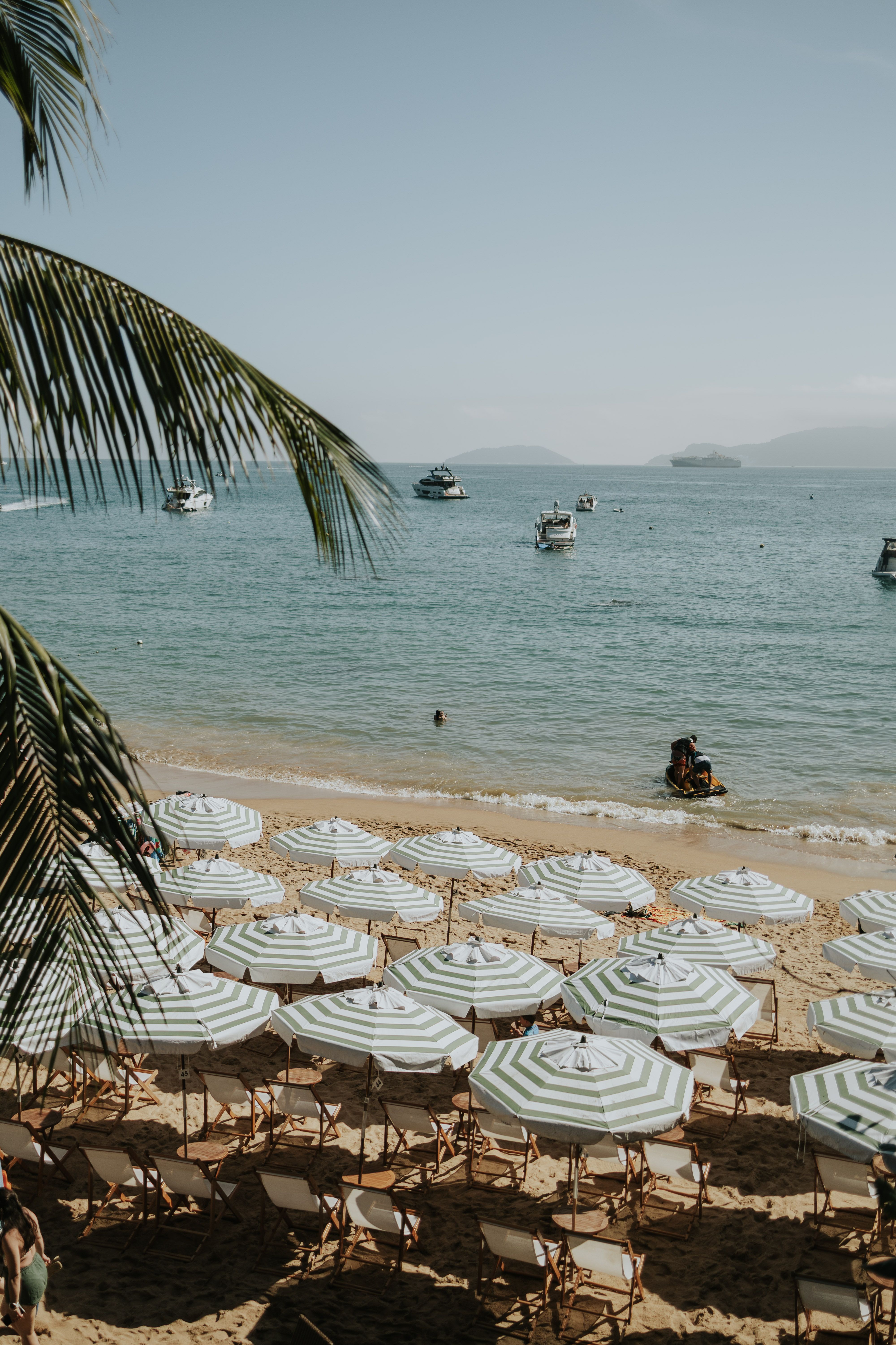 A tranquil beach scene with striped umbrellas and boats on a sunny, clear day.