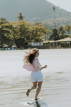 A woman in a pink shirt joyfully runs on a tropical beach with scenic mountains.