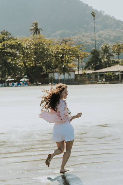 A woman in a pink shirt joyfully runs on a tropical beach with scenic mountains.