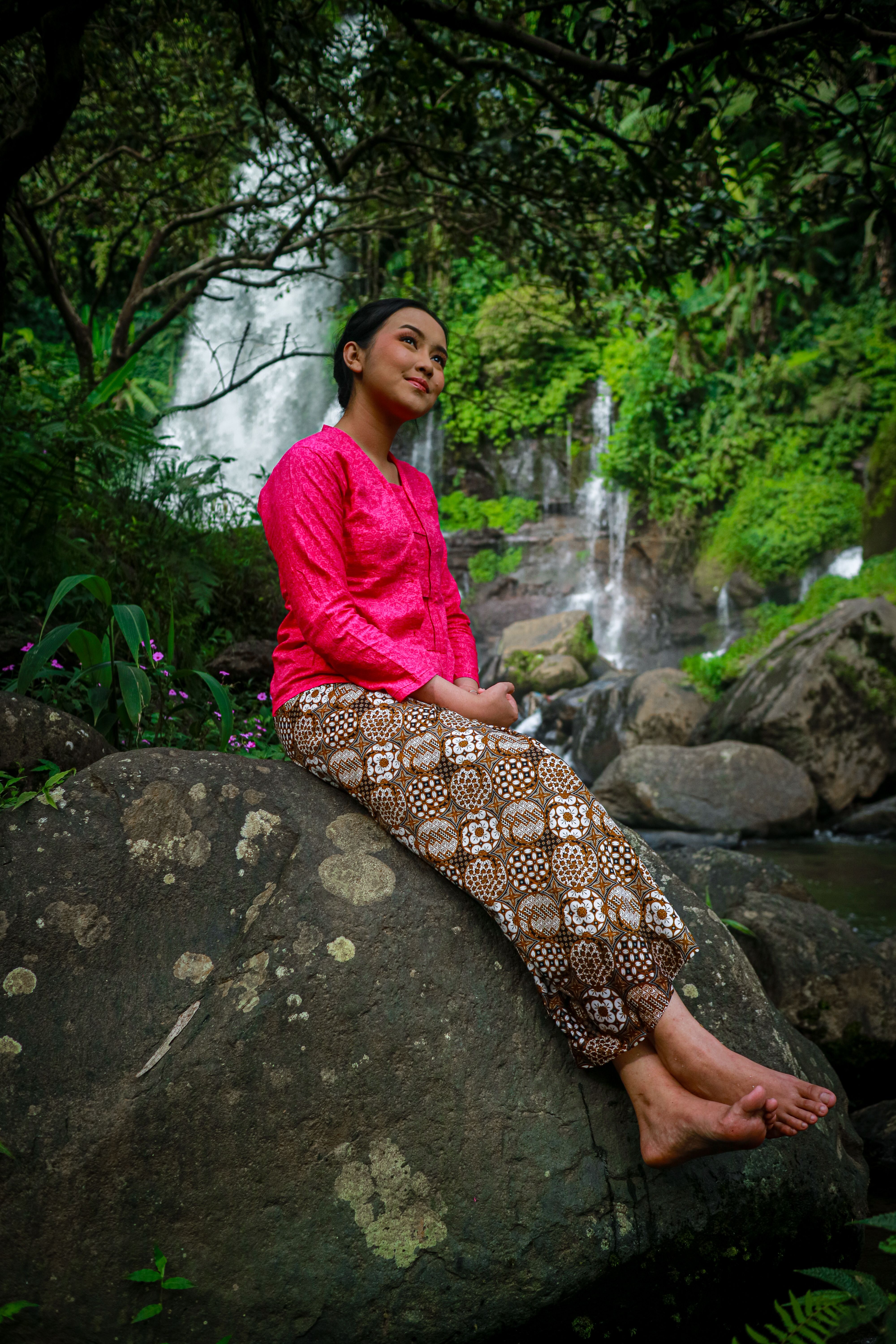 A serene portrait of a woman in traditional attire sitting by a waterfall in a lush forest in Indonesia.