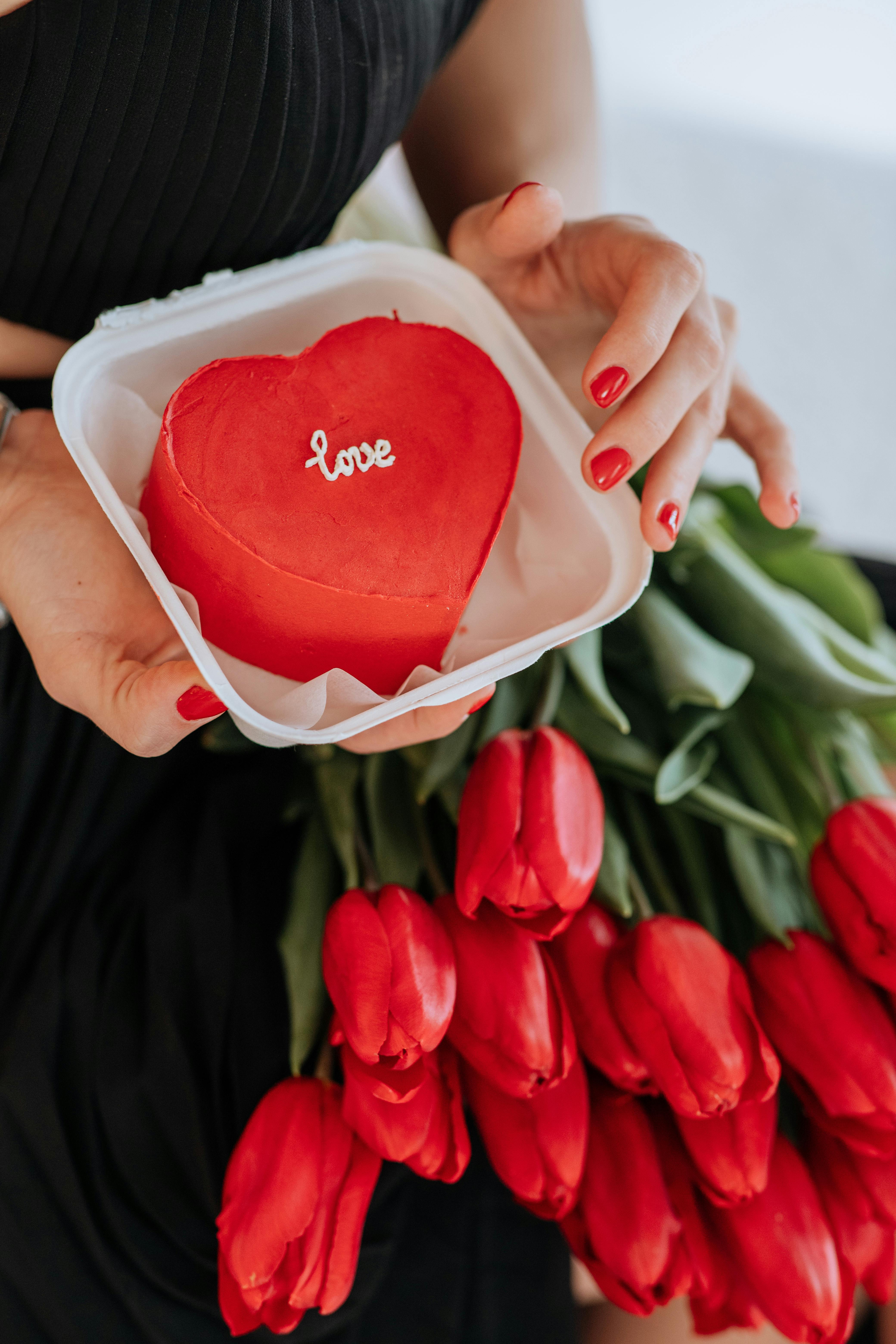 Heart-shaped red cake with 'love' text and bouquet of tulips symbolize romance.