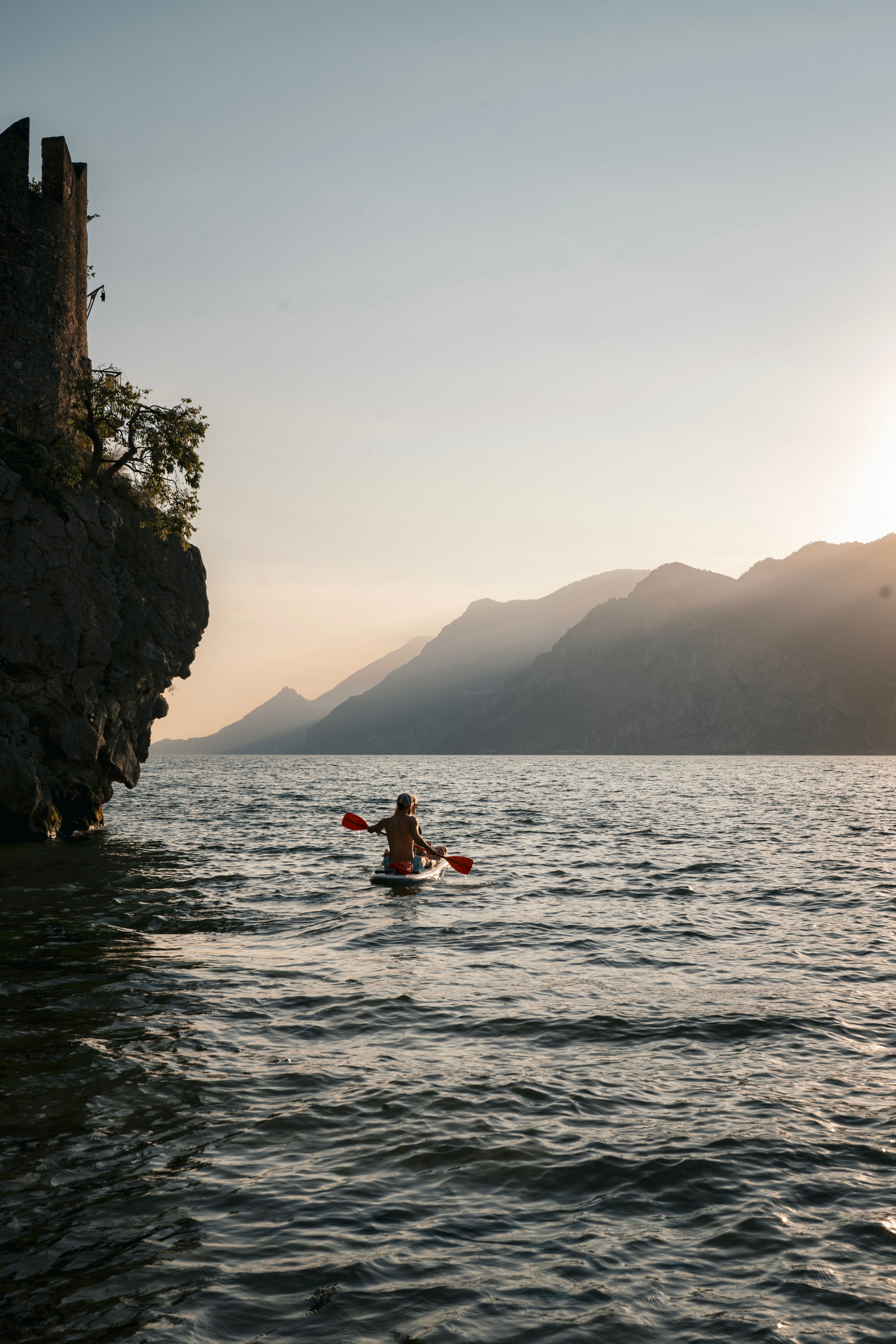 A lone kayaker paddles on Lake Garda with a scenic view of the mountains at sunset in Malcesine, Italy.