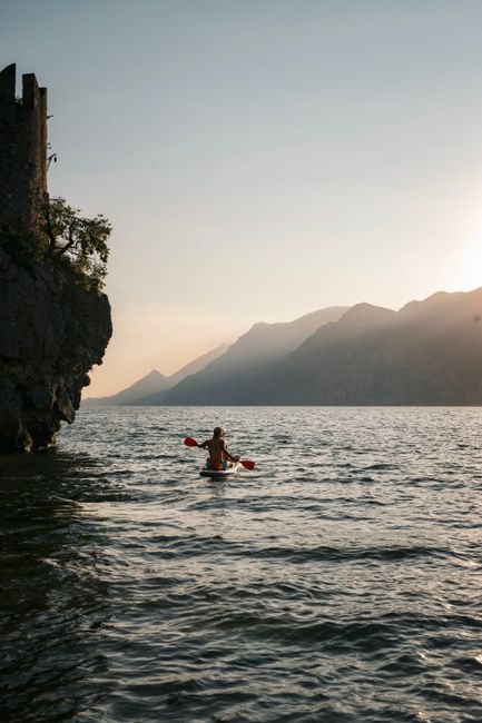 A lone kayaker paddles on Lake Garda with a scenic view of the mountains at sunset in Malcesine, Italy.