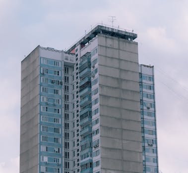 A tall residential building with multiple balconies set against a cloudy sky, showcasing urban architecture.