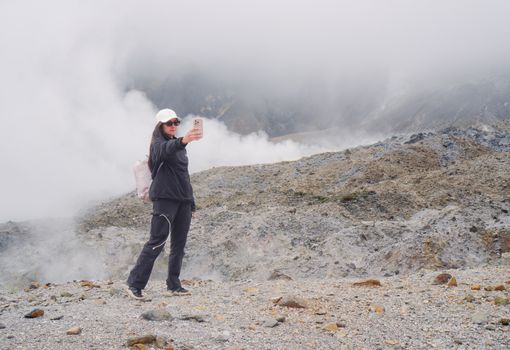 A woman captures a selfie on her phone at a foggy volcanic landscape, showcasing outdoor exploration.