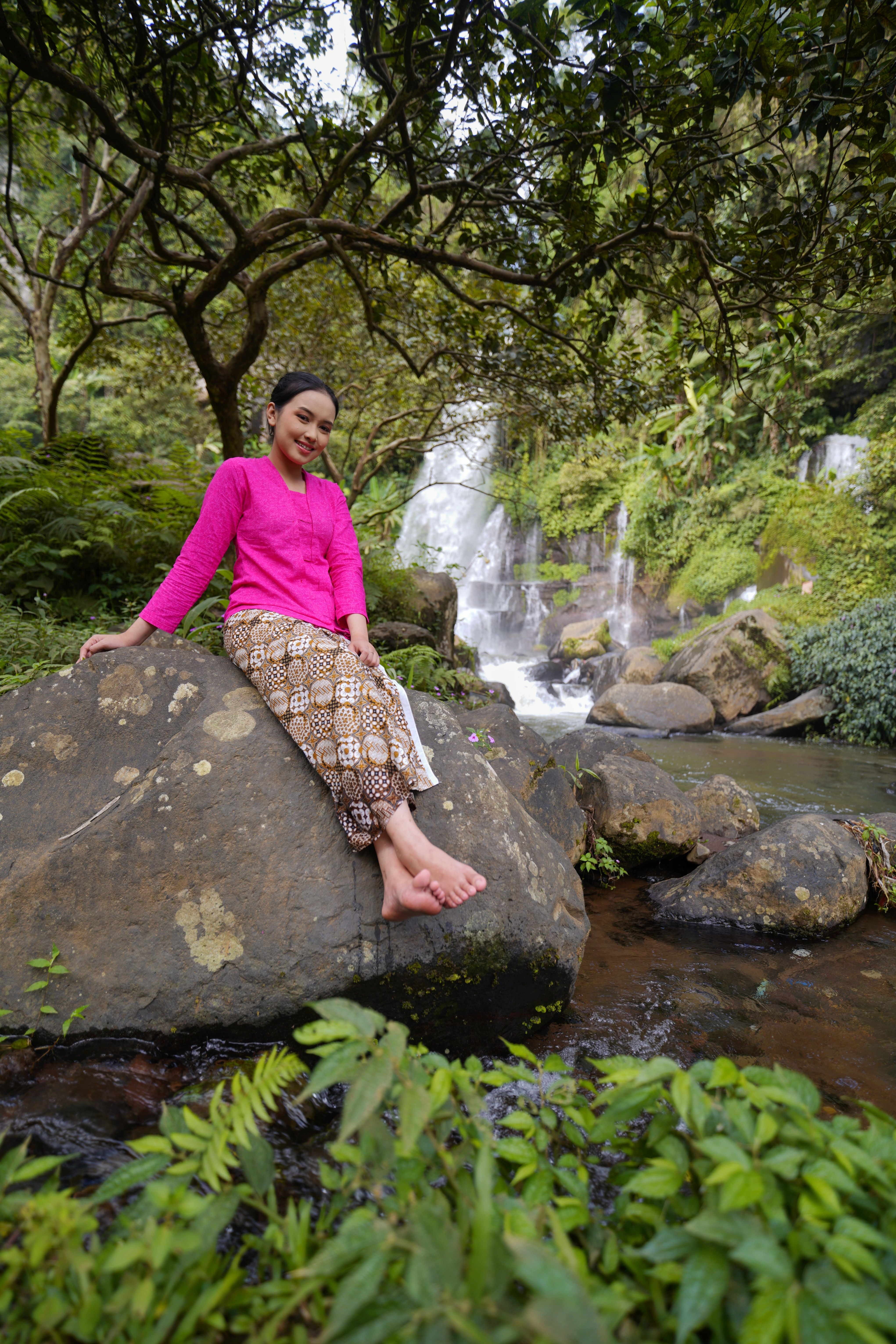 A woman in traditional attire sits gracefully on a rock by a scenic waterfall in Jawa Barat, Indonesia.