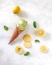 Top view composition of ice cream in waffle cone places near fresh lemon and lemon slices with scattered between green mint leaves on marble surface