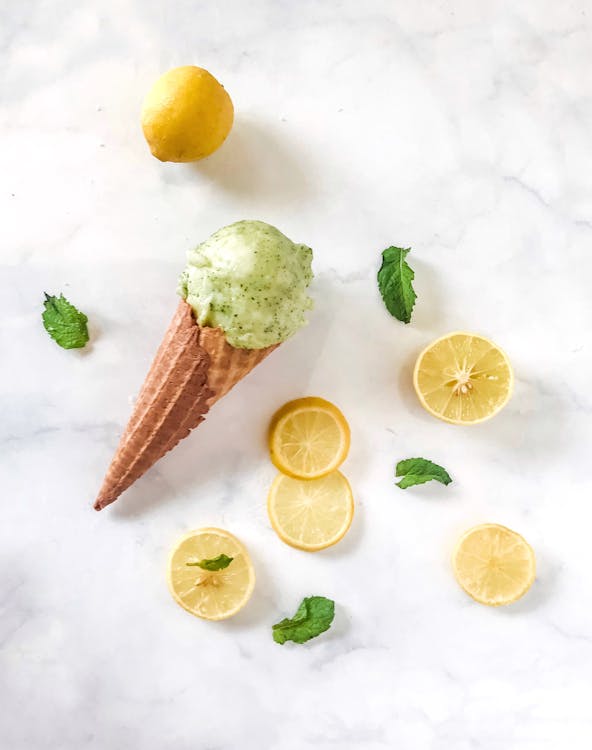 Top view composition of ice cream in waffle cone places near fresh lemon and lemon slices with scattered between green mint leaves on marble surface