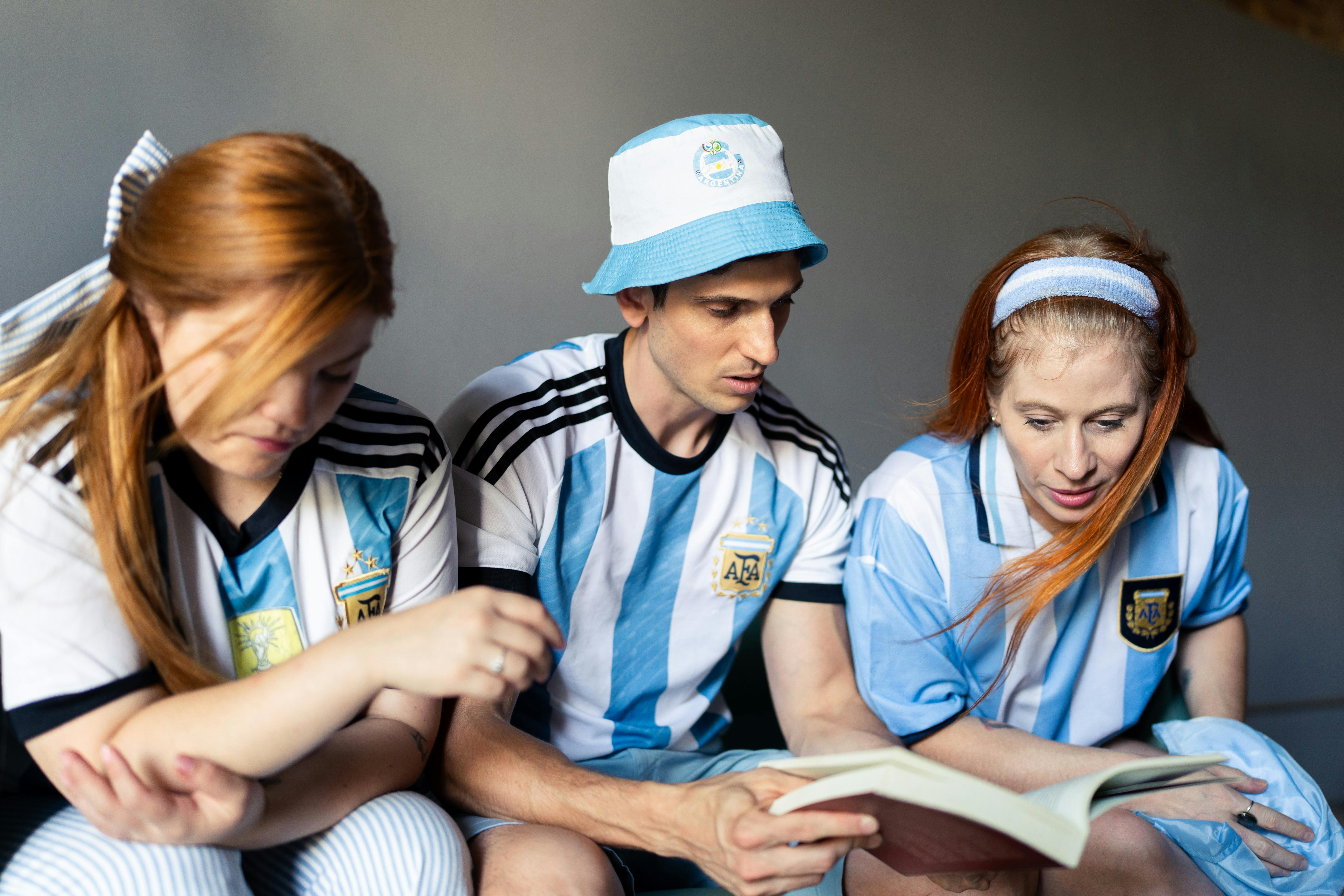 Three fans in Argentina jerseys engage with a book, showing team spirit indoors.
