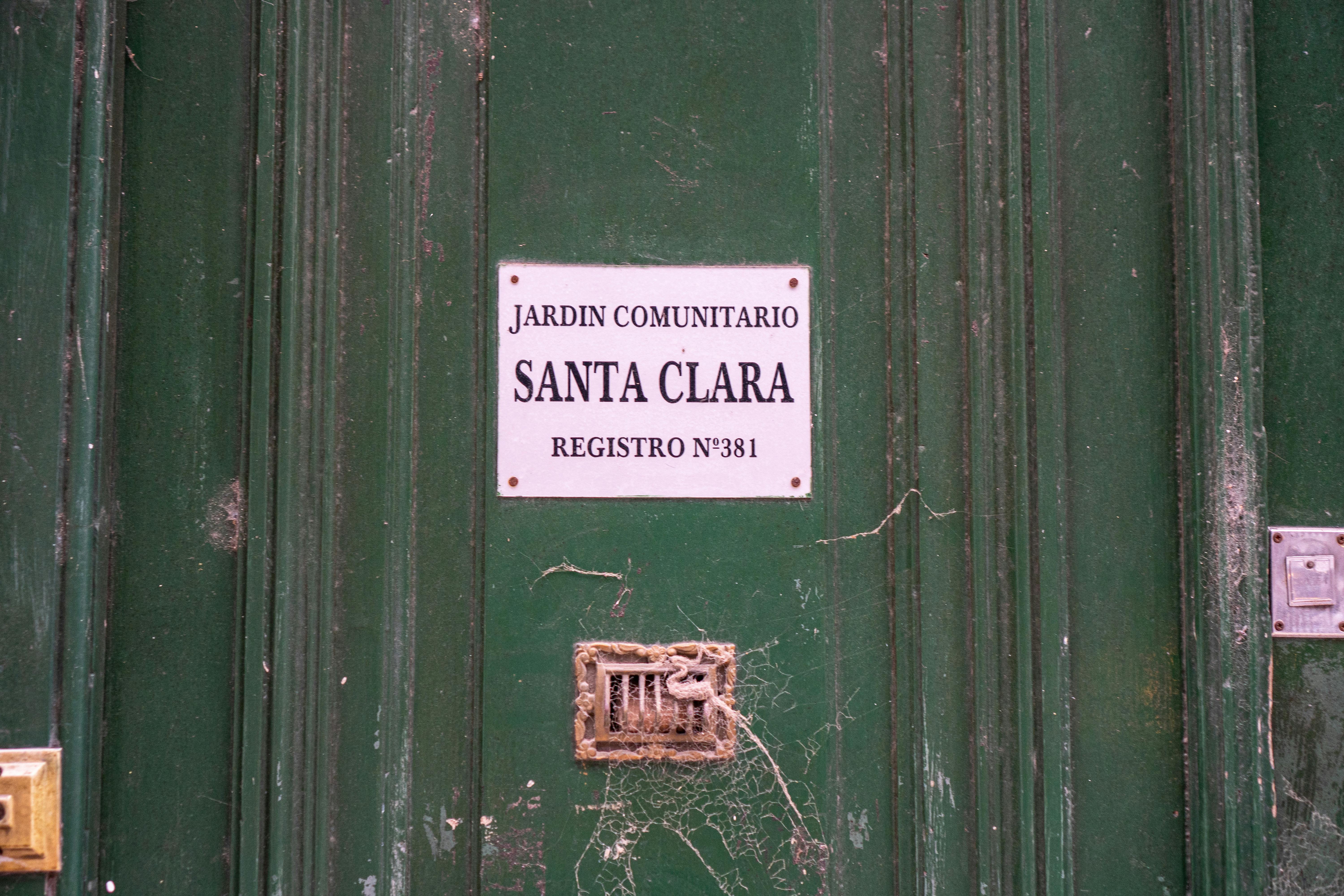 grátis Porta verde desgastada com placa indicando o jardim comunitário 'Santa Clara' e teias de aranha. Foto profissional