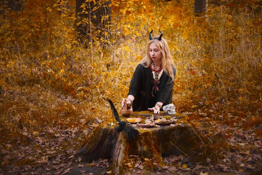 A witch in horns performs a ritual in an autumn forest with leaves and mystical items.