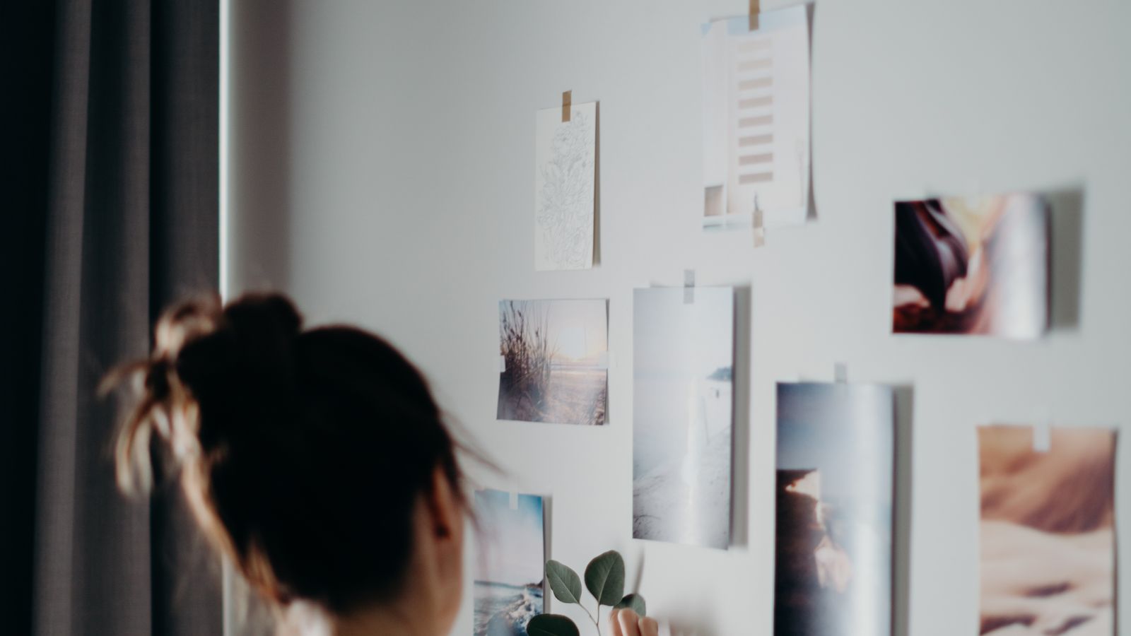 A woman arranging printed photographs on a white wall indoors