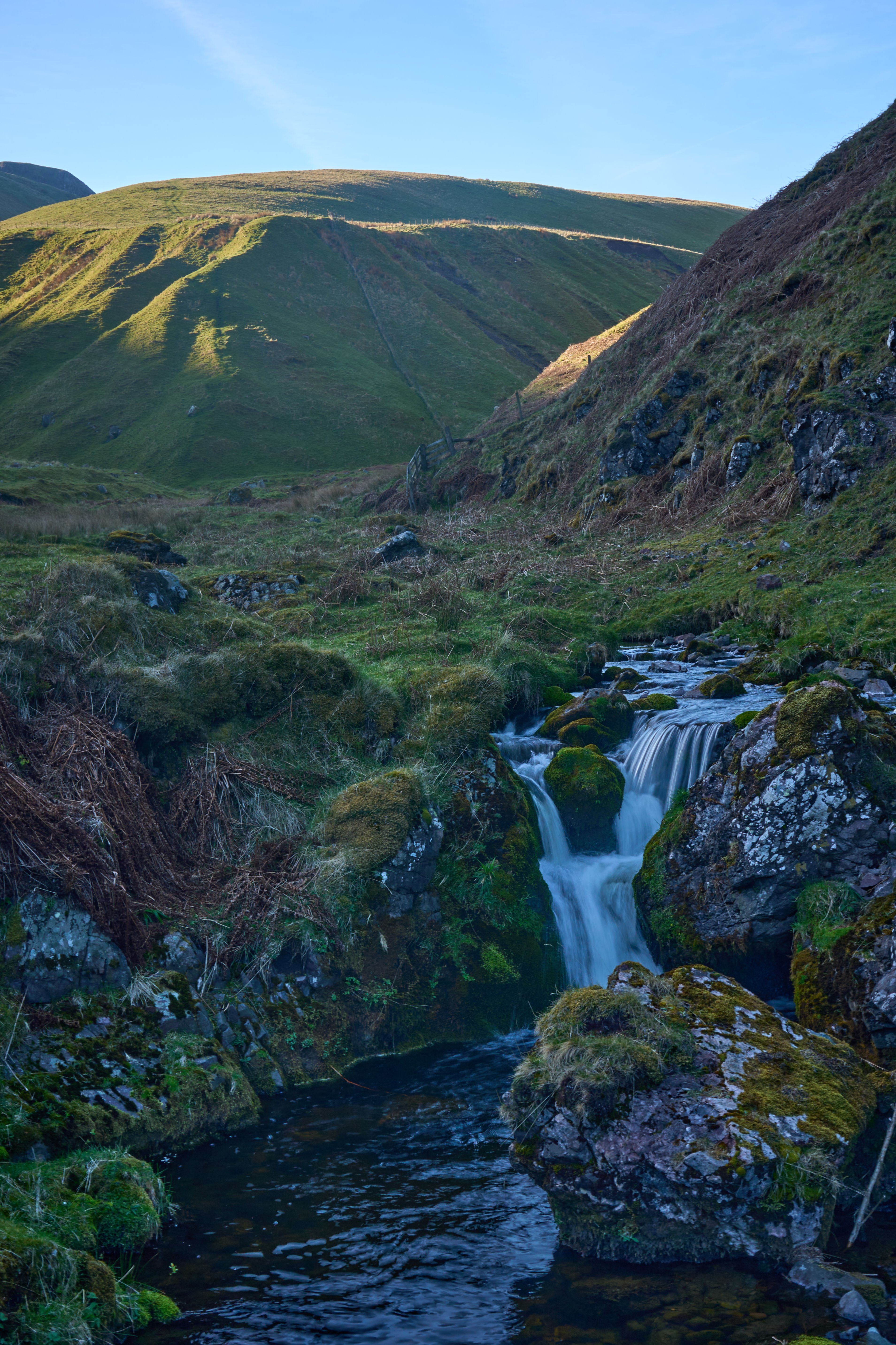 Gratuit Une cascade sereine coule à travers les Highlands écossaises moussues, entourée de collines verdoyantes baignées de soleil. Photos