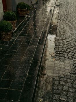 Wet cobblestone street in Bruges, Belgium, with wicker baskets and bushes reflecting in rain puddles.