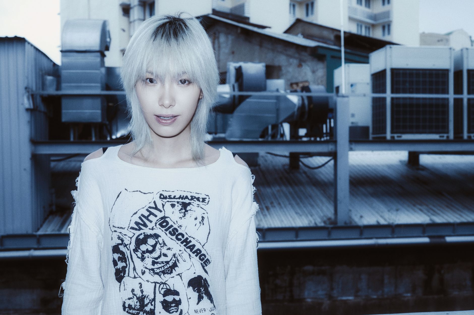 Stylish young adult with platinum hair stands against urban industrial backdrop.