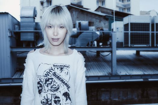 Stylish young adult with platinum hair stands against urban industrial backdrop.