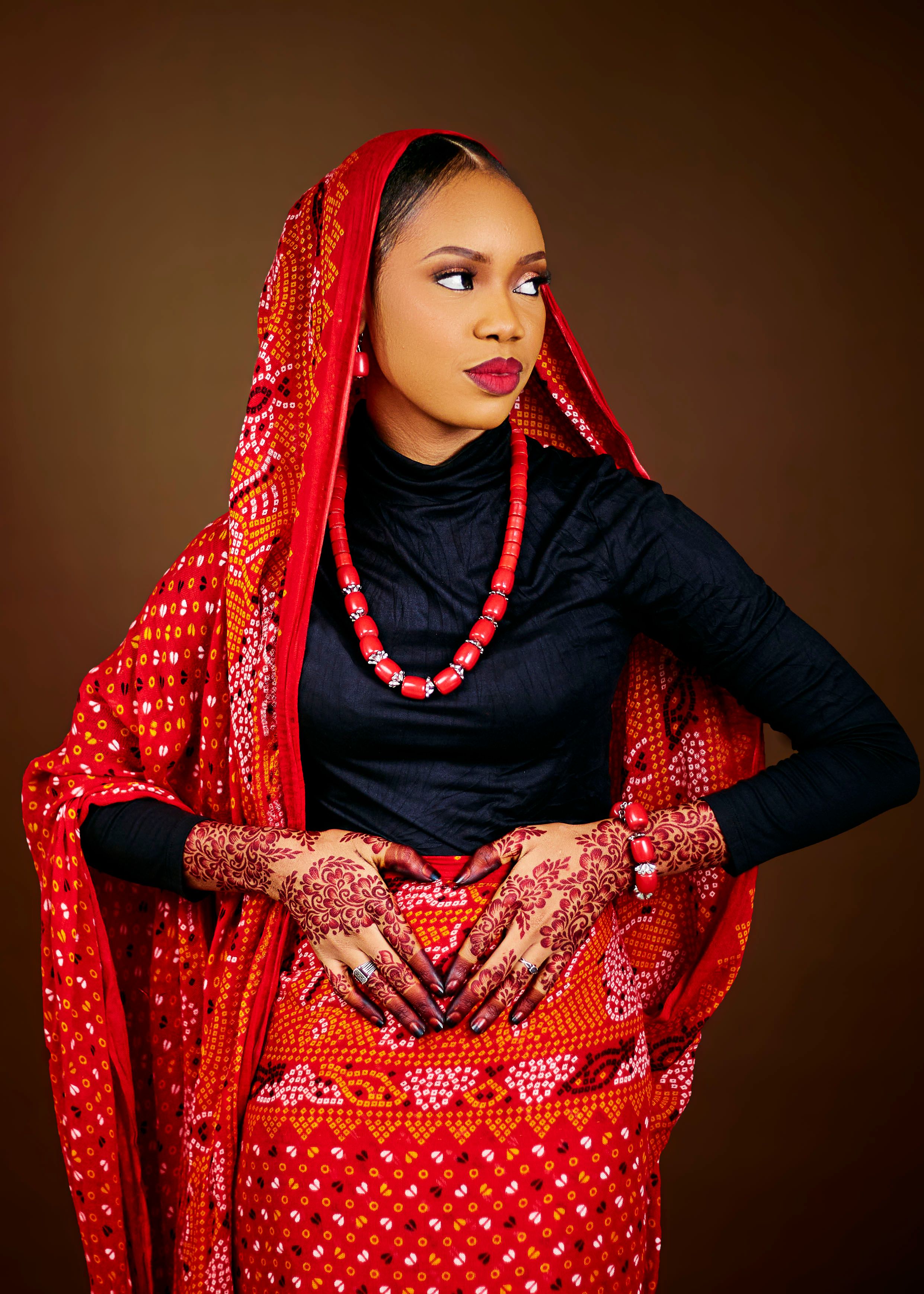 A woman wearing red traditional attire with henna decorated hands against a brown background.