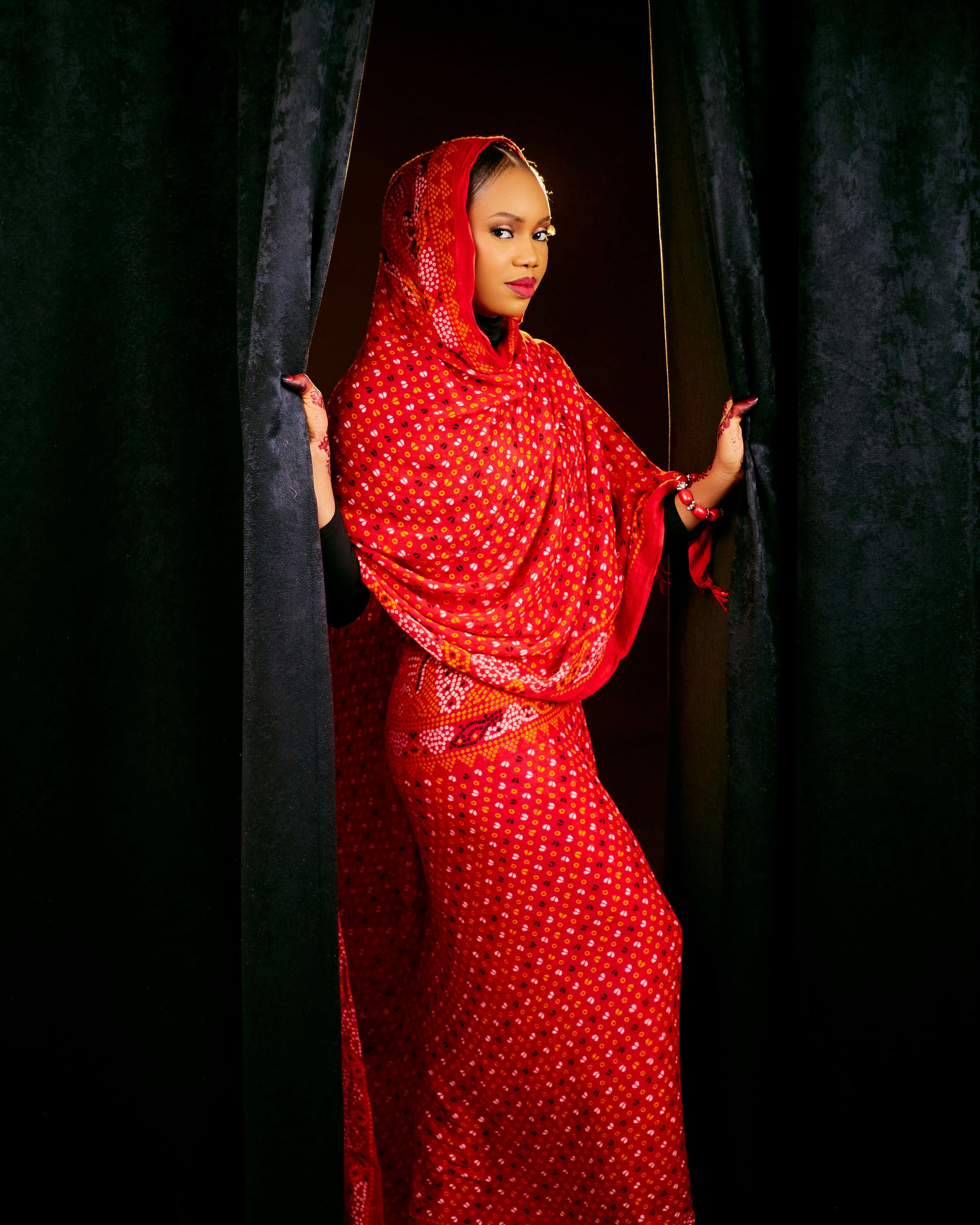 A woman in beautiful traditional red attire poses gracefully between curtains.