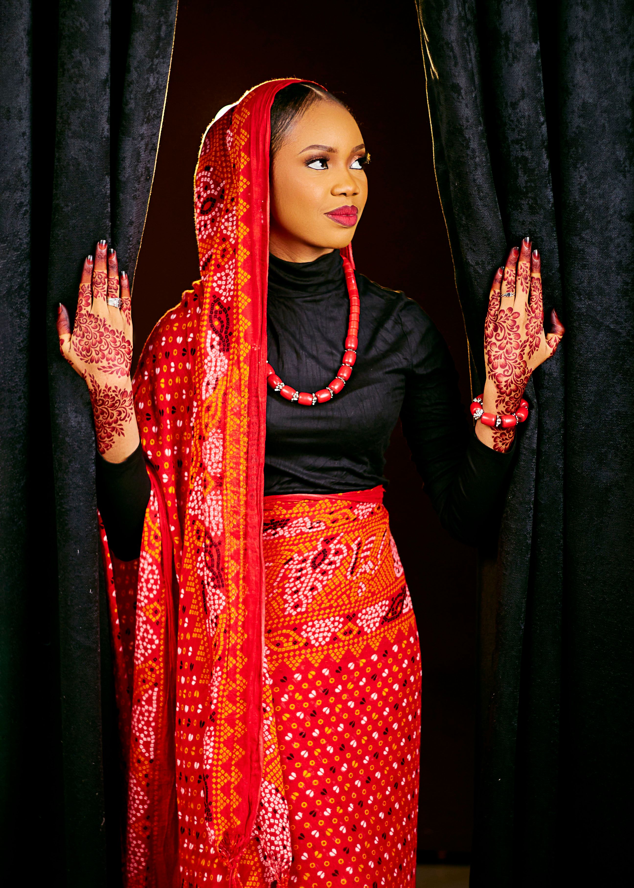 A woman in vibrant traditional attire holding curtains, showcasing intricate henna designs.