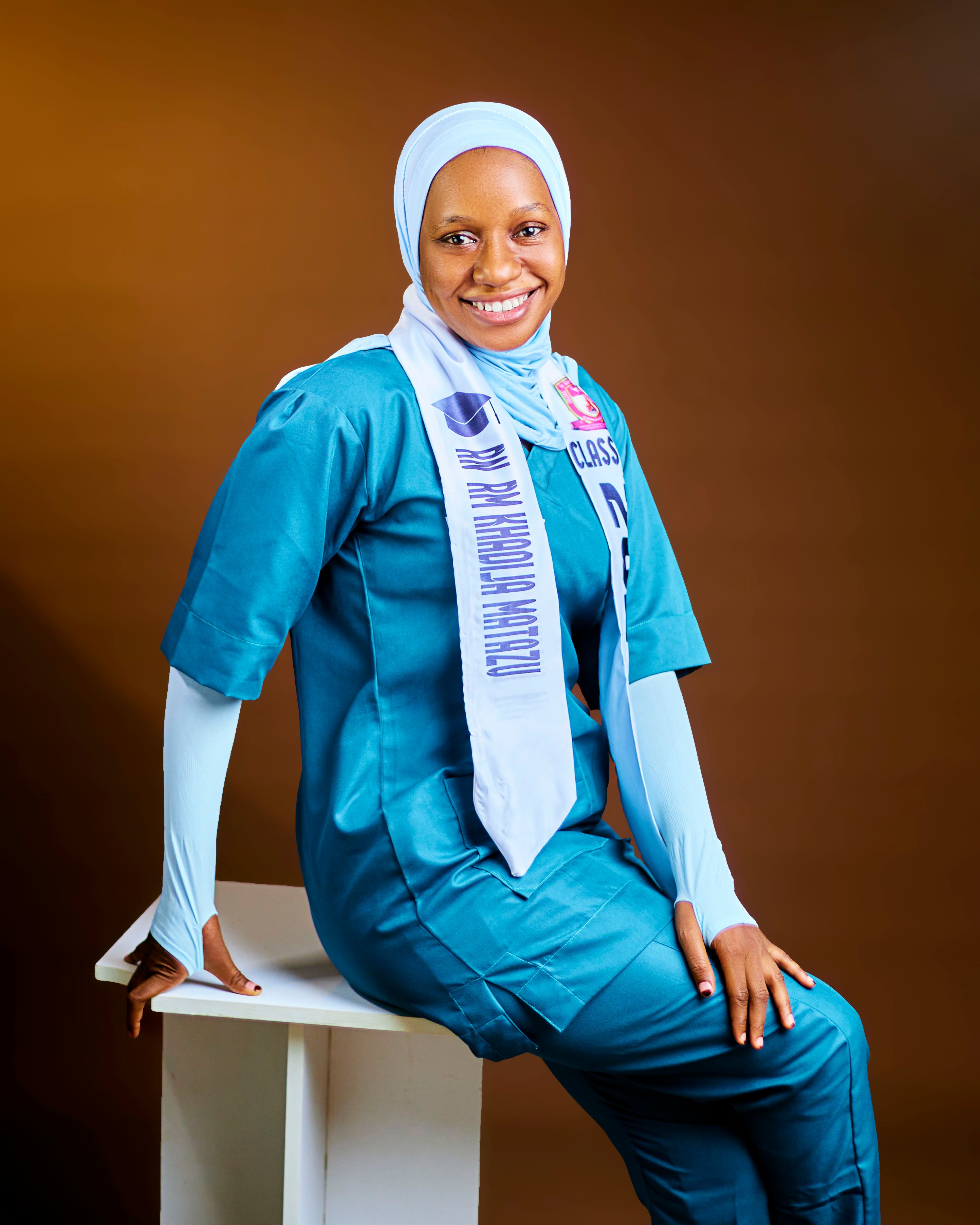 A nurse in scrubs and hijab, seated and smiling with a graduation stole.