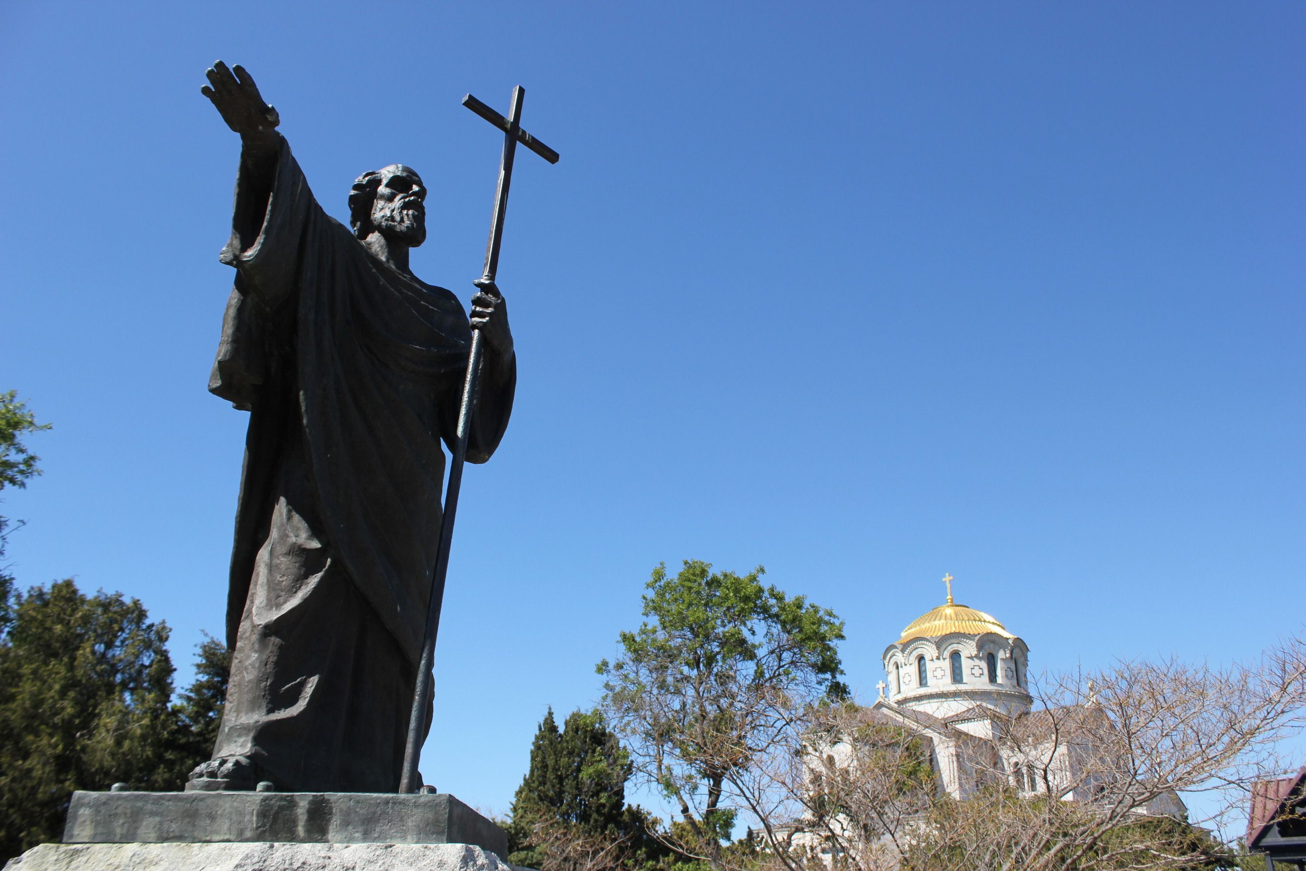 A bronze statue of a saint holding a cross, with a church dome under a clear blue sky.