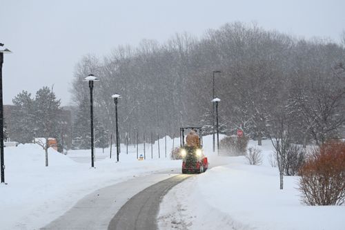 Free A snow plowing tractor clearing a snowy path lined with trees and street lamps. Stock Photo