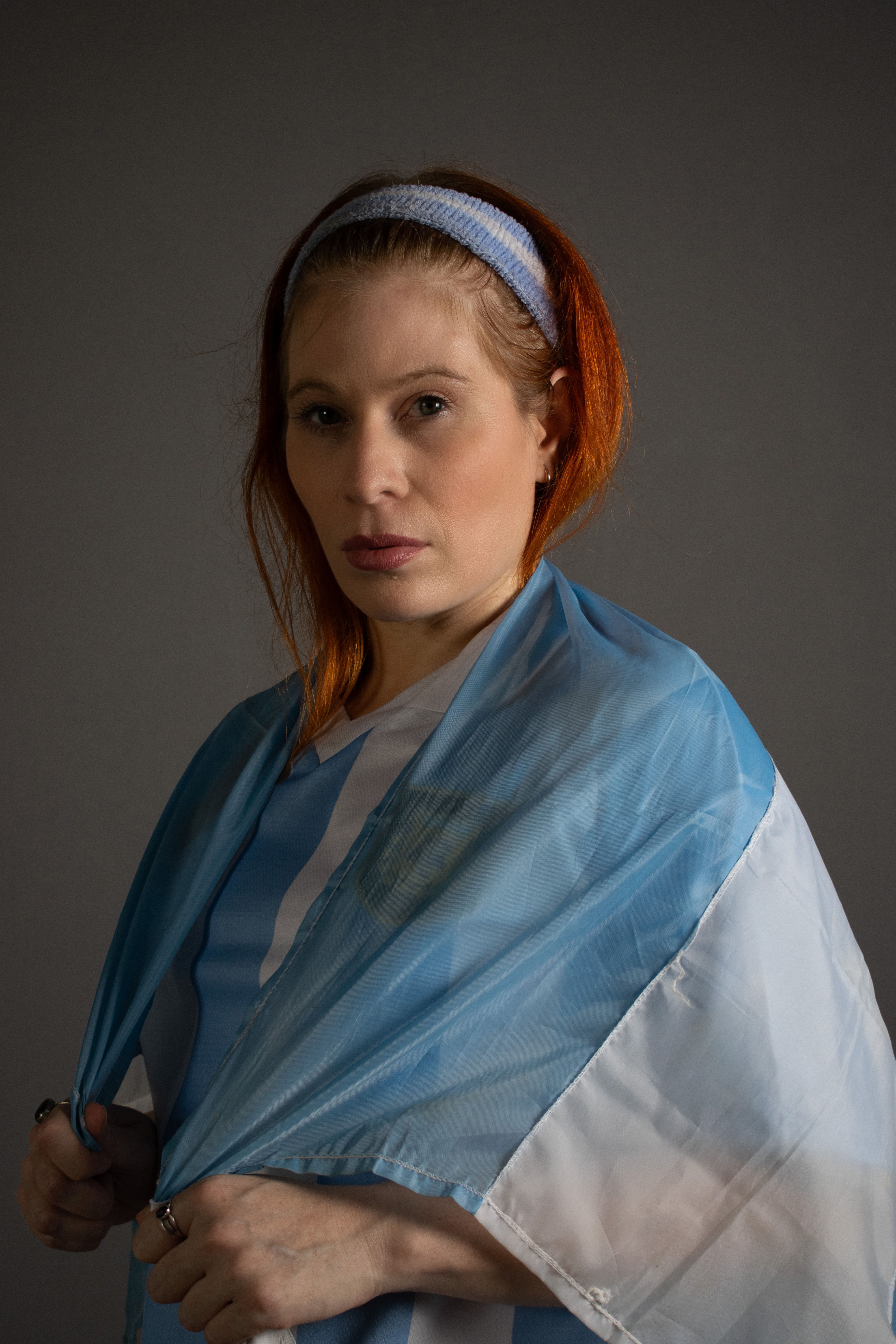 Portrait of a woman with red hair draped in fabric, looking confident and elegant against a neutral background.