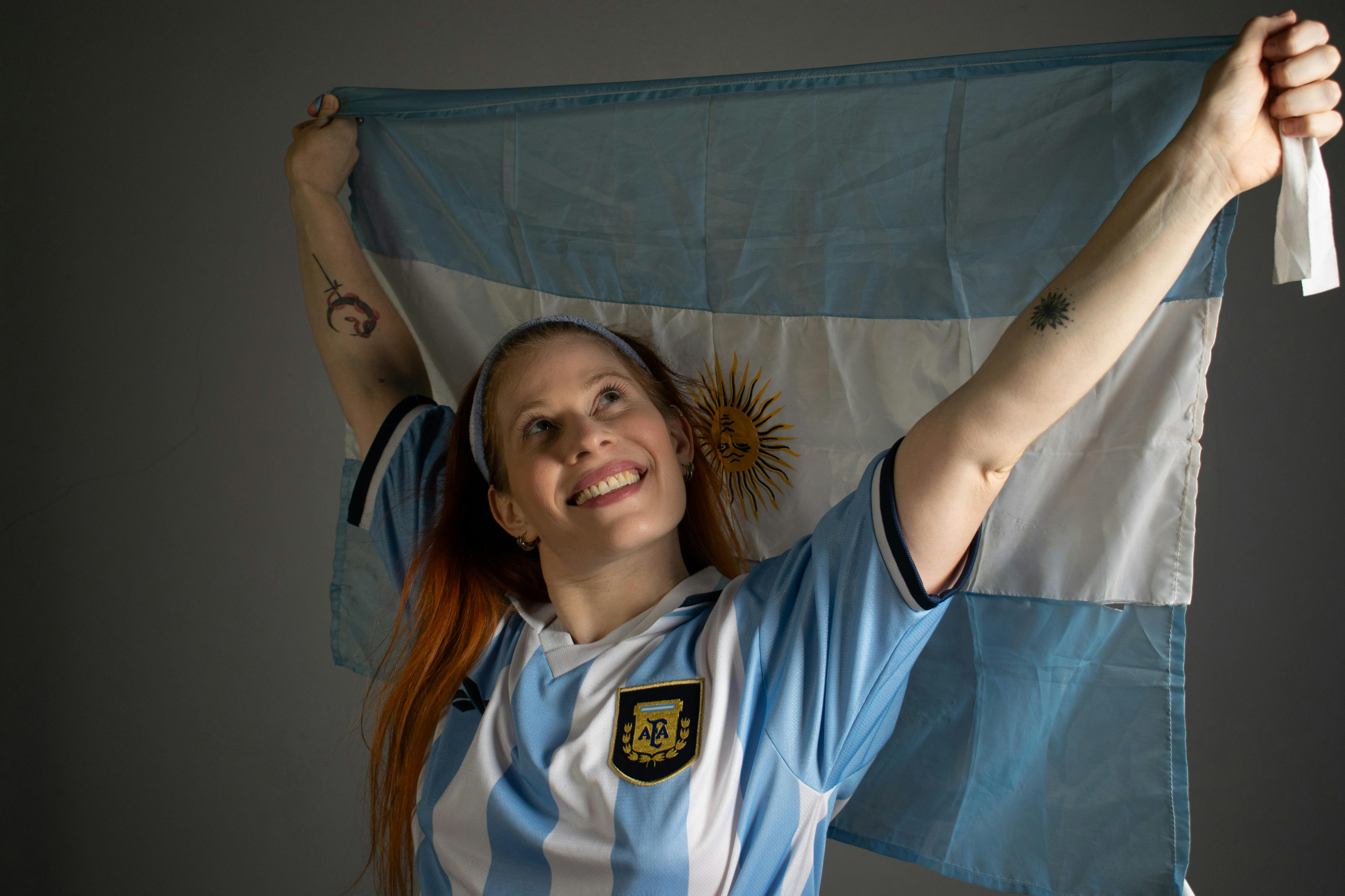 Smiling woman holding up the Argentina flag in celebration, wearing a blue and white jersey.