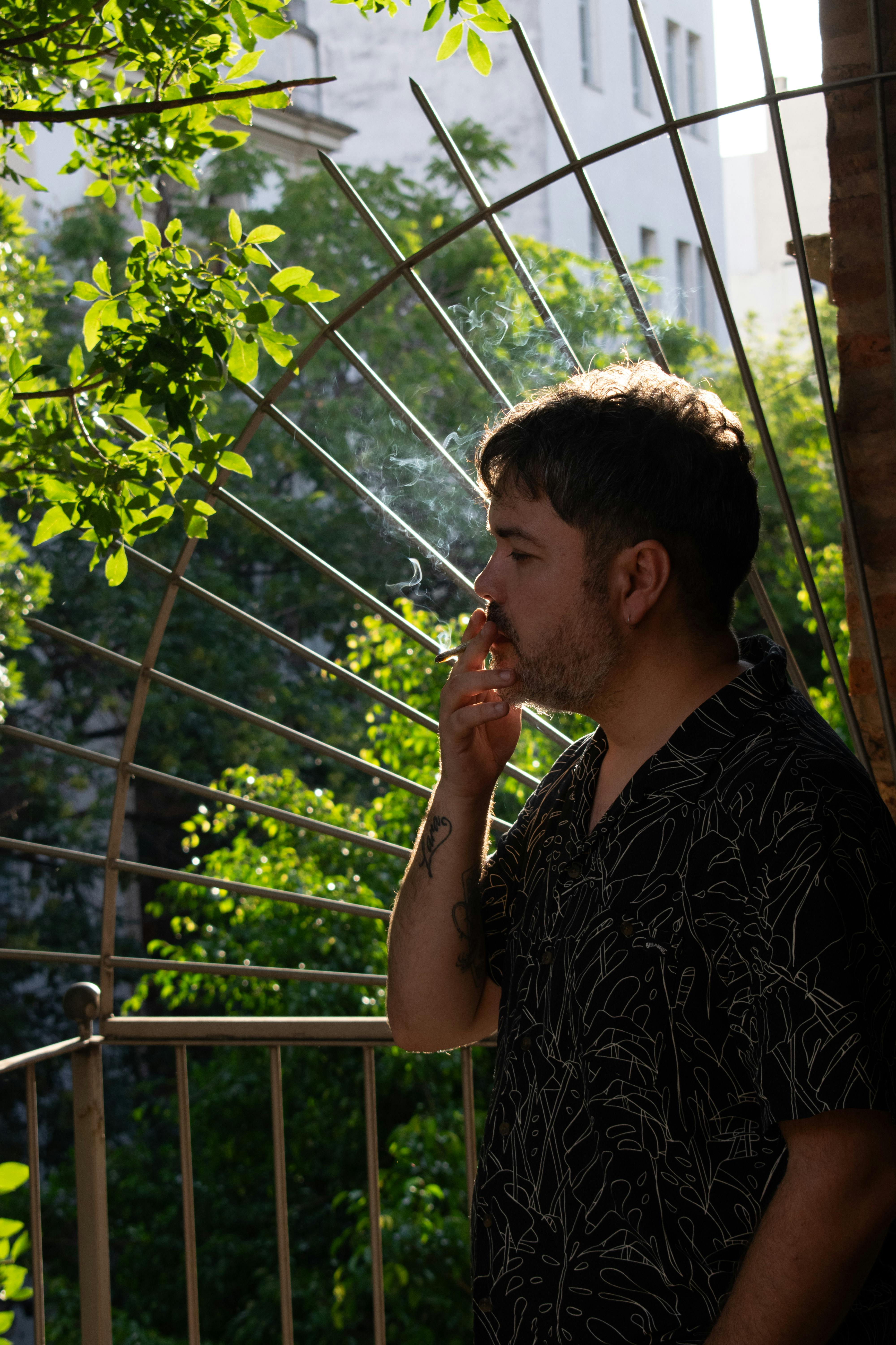 A man smoking a cigarette on a balcony with sunlight filtering through leaves.