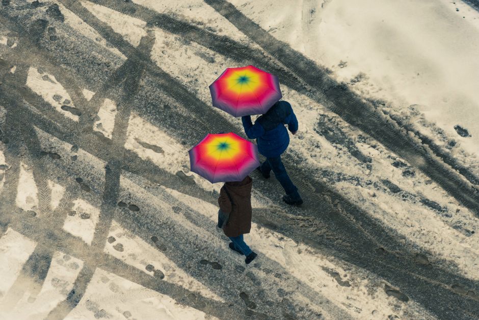 Two people walk on a snowy street with vibrant umbrellas, viewed from above.