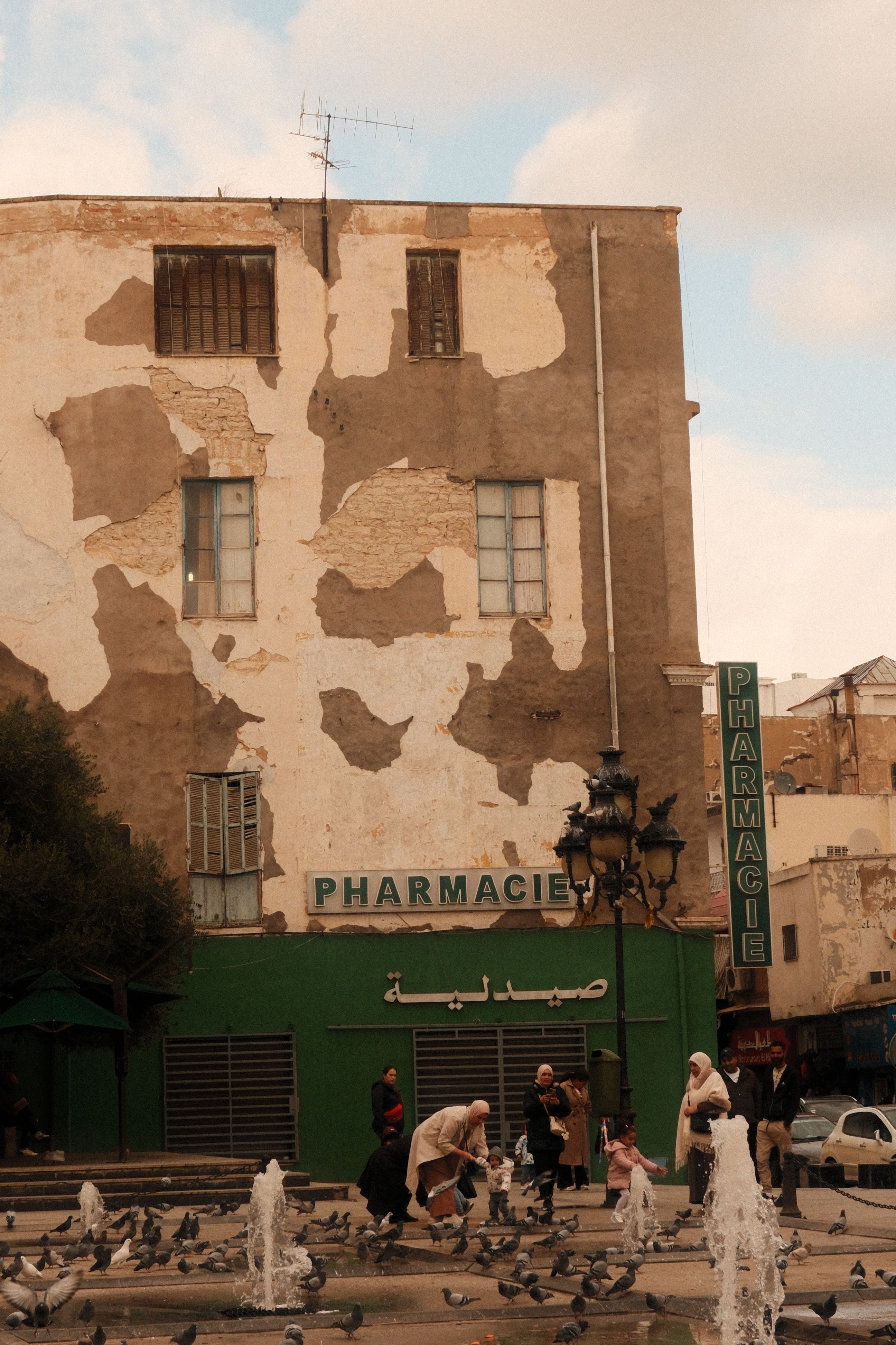 Exterior view of a Tunisian pharmacy with people and pigeons in front, showcasing local architecture.