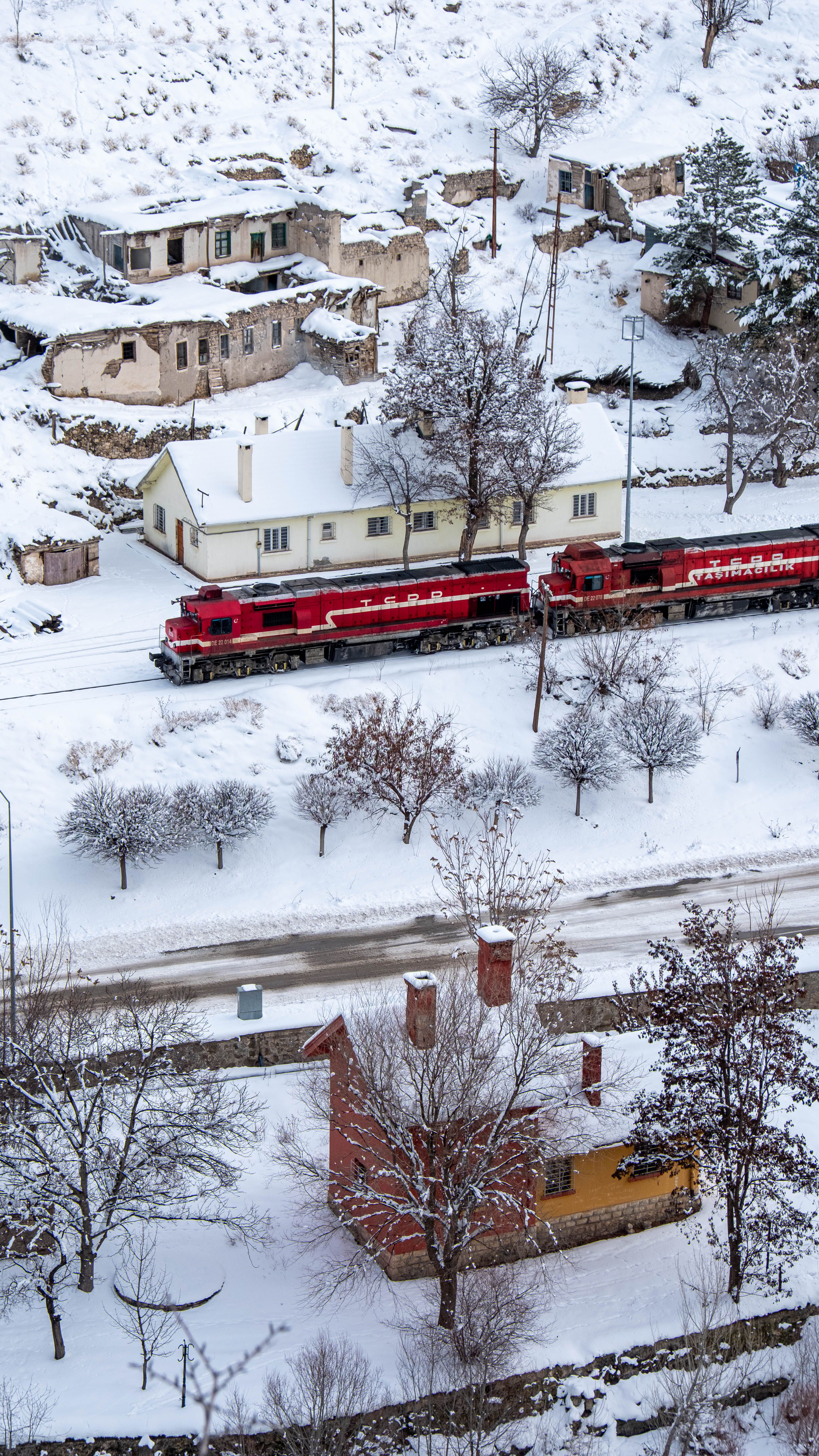 Red train travels through a snow-covered village in a winter wonderland scene.