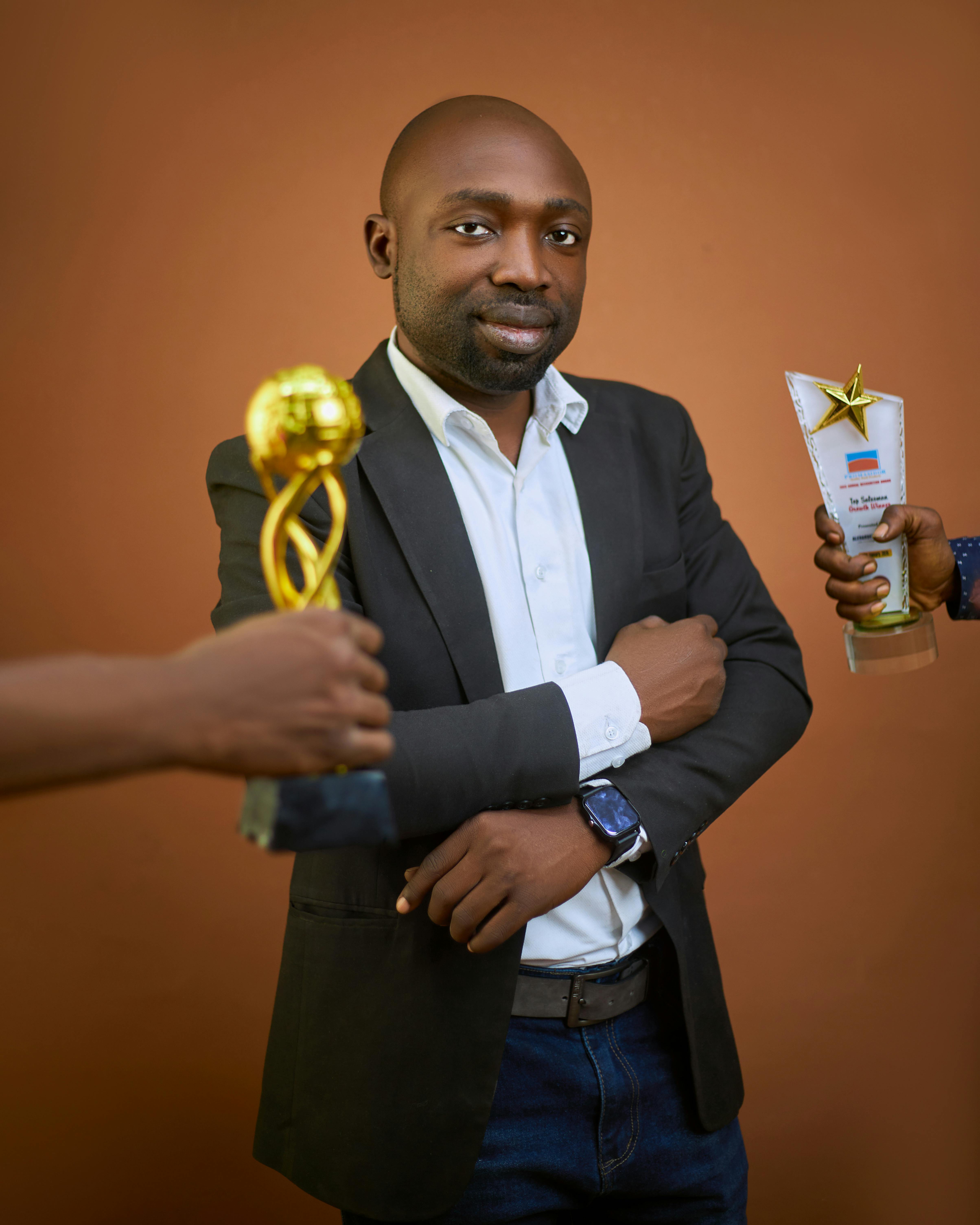 Confident man in a suit holding awards and trophies against a neutral background.