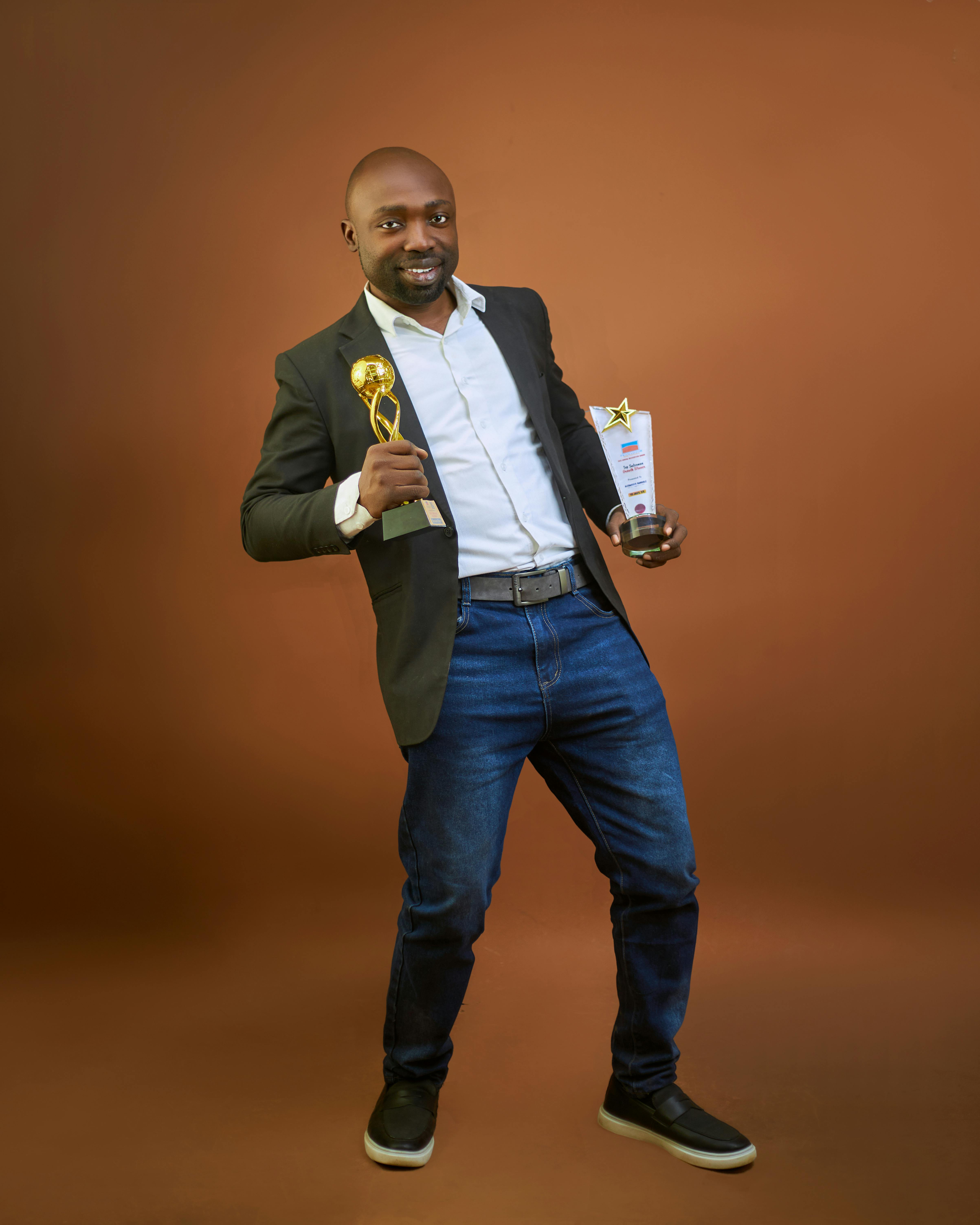 grátis Homem alegre de terno segurando dois troféus contra um fundo marrom. Foto profissional