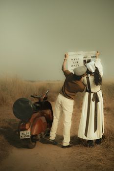 Couple in vintage attire with a scooter holding a 'Save the Date' sign in a field.