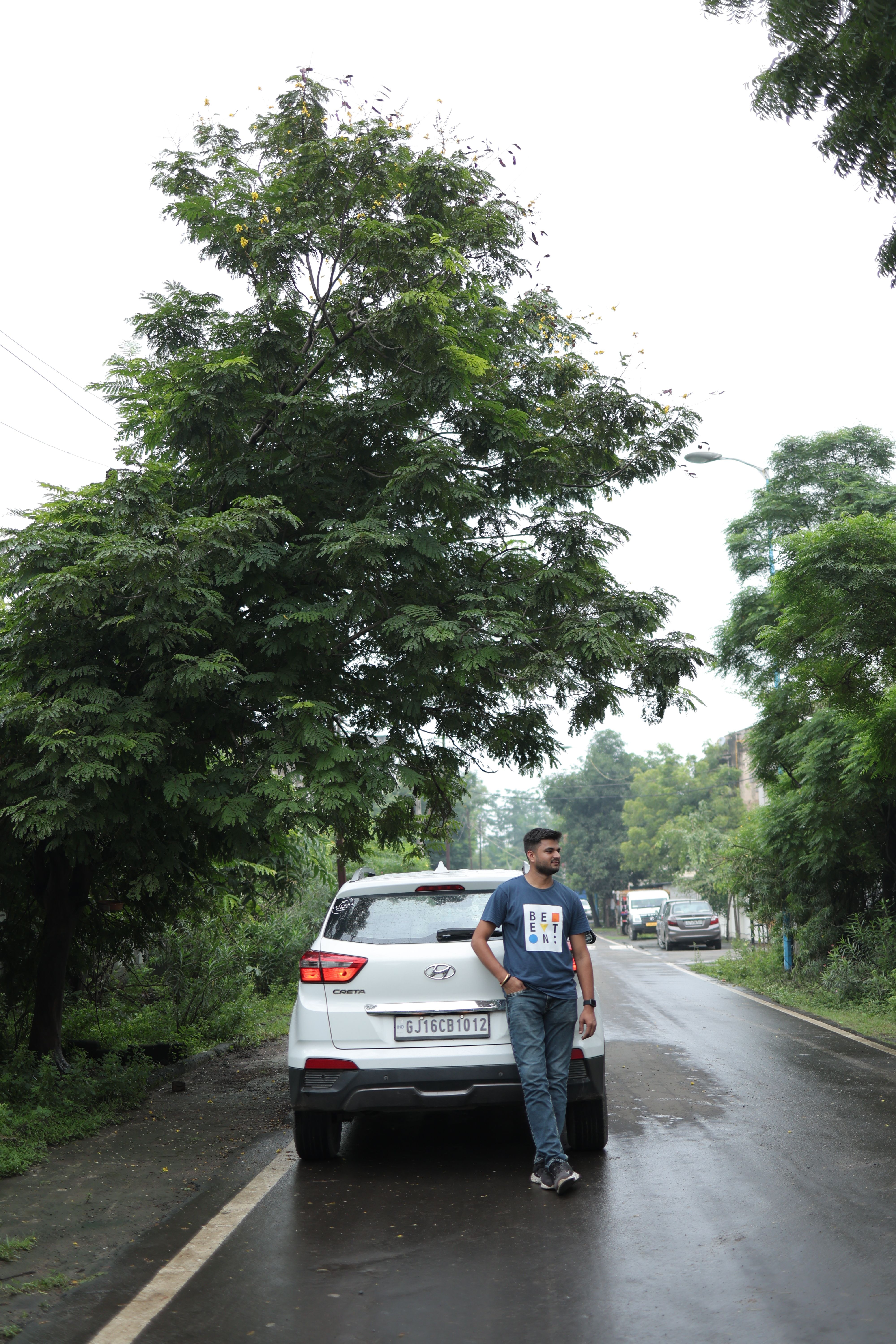 A man stands leaning against a car on a wet road, surrounded by lush greenery.