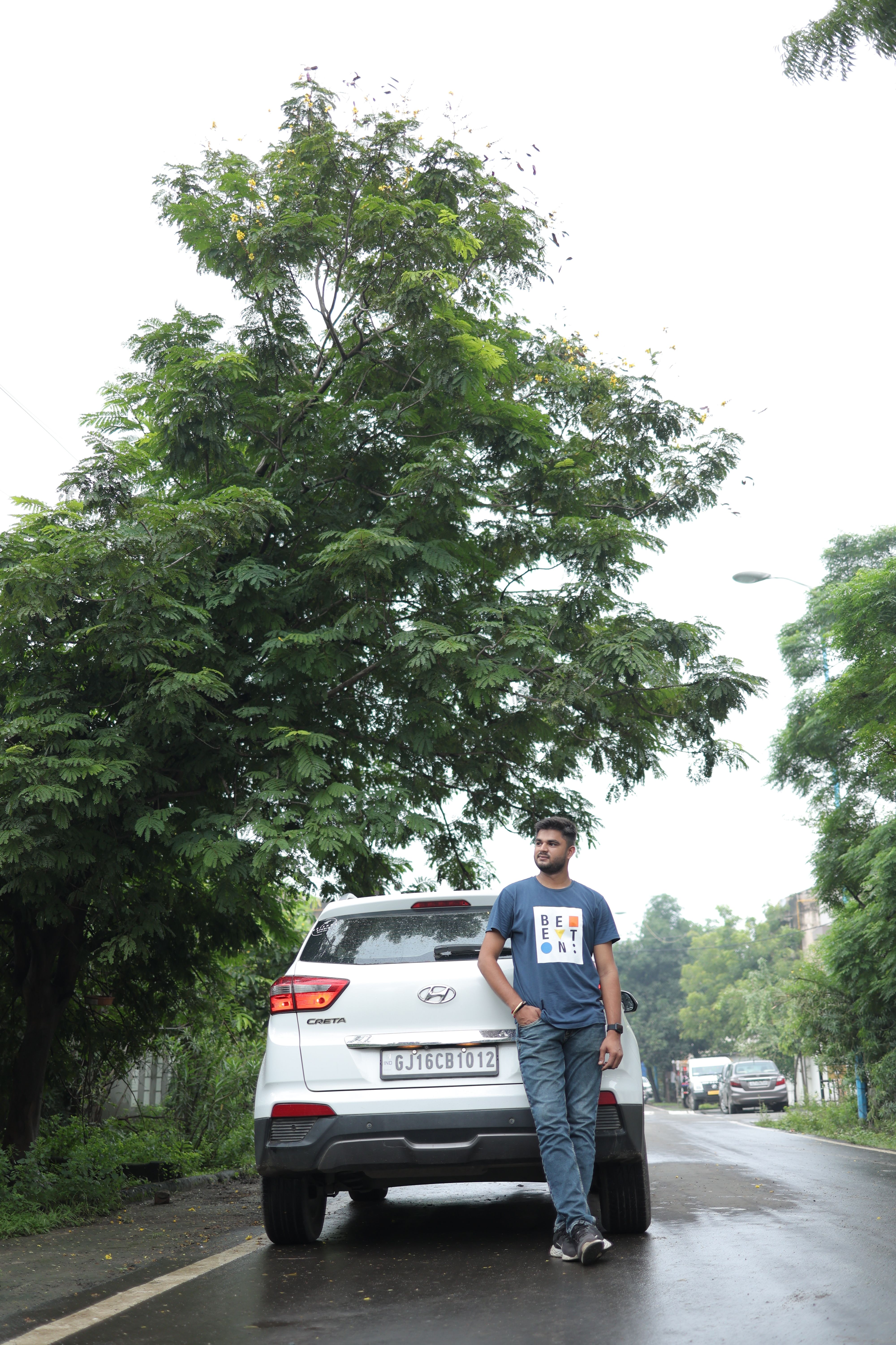 Man casually posing by a white car on a serene, tree-lined road, showcasing modern lifestyle.